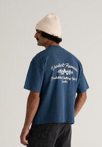 Mies, joka pukeutuu siniseen t-paitaan, jossa on kukkateksti "Verdant Reverie Prohibited Clothing USA Berlin" selässä sekä kermaiseen neulottuun pipoon.