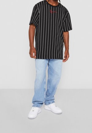 Black and white striped t-shirt with red emblem, paired with light blue jeans and white sneakers. Casual fit with short sleeves.