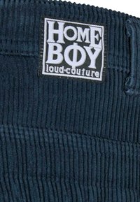 Tumši zils kordurojs ar izteiktām vertikālām līnijām; satur baltu etiķeti ar "HOMEBOY loud.couture" logo. Redzama teksturēta auduma detaļa.