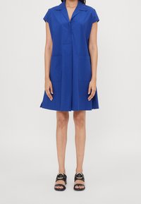 Donna che indossa un vestito blu a maniche corte, lungo fino al ginocchio, con tasche frontali e sandali neri aperti alla punta con fibbie dorate.