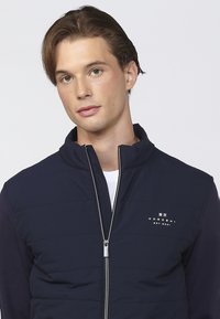 Marineblå lynlåsjakke med quiltet front, høj krave og lange ærmer. Har logo på brystet og metal lynlås. Glat stofstruktur.