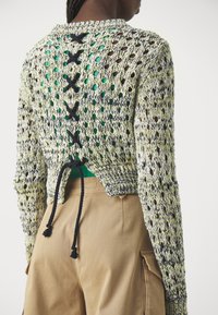 Osoba nosząca sweter z dzianiny o otwartej strukturze, z czarnymi krzyżującymi się sznurkami na plecach, na zielonej bluzce i beżowych spodniach cargo.