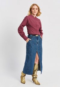 Donna che indossa un top a maniche lunghe bordeaux, una gonna di jeans a vita alta con spacco frontale, fibbia della cintura dorata e stivali dorati morbidi, con le mani nelle tasche.