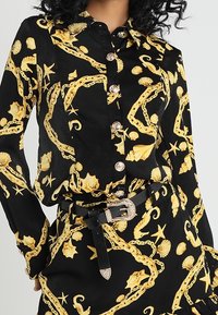 Svart långärmad skjorta och kjol med gula snäcks- och kedjemönster, knäppt framtill, samt svart bälte med silverspänne.