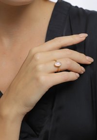 Bague en or ornée d'une pierre rose ovale. L'anneau est lisse, minimaliste, et repose sur une main de couleur claire sur un fond en tissu sombre.