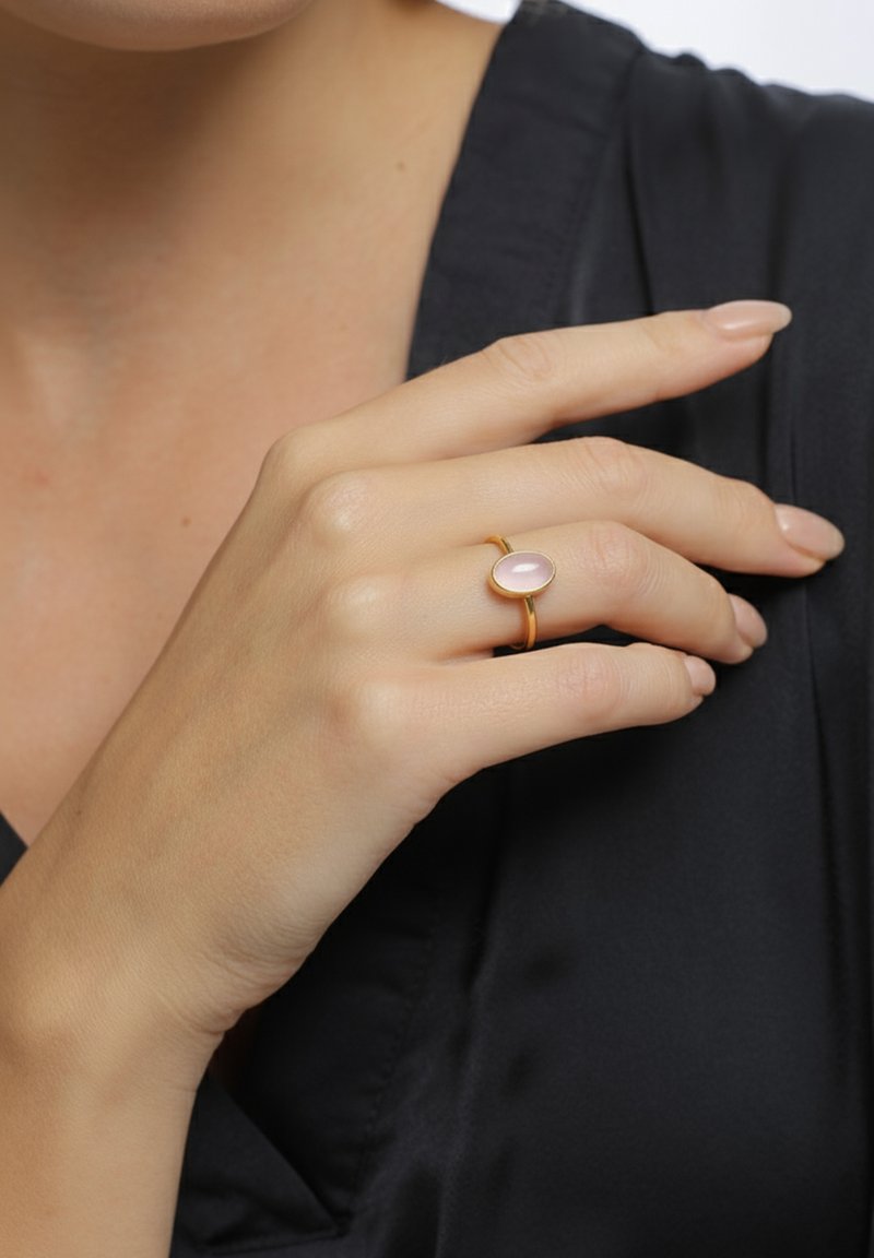 Bague en or ornée d'une pierre rose ovale. L'anneau est lisse, minimaliste, et repose sur une main de couleur claire sur un fond en tissu sombre.