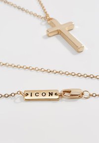 Gulltone halskjede med en elegant korspendant og en rektangulær plakk som leser "IKON." Har en tynn kjede og en klipslås.