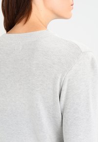 Jasnoszary sweter z żebrowanym dekoltem i fakturą, charakteryzujący się bezszwowymi detalami, widziany z tyłu, ukazujący szczegóły na ramionach.