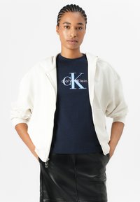 Felpa crema con zip sopra una t-shirt navy con logo Calvin Klein, abbinata a una gonna di pelle nera. Texture lisce e colori a contrasto.