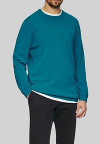 Teal Langarm-Sweatshirt mit Rundhalsausschnitt, gerippten Bündchen und Saum. Über einem weißen Shirt getragen, kombiniert mit schwarzen Hosen.