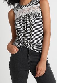 Czarny i biały paskowany top bez rękawów z koronkowym panelem, zebranym projektem i węzłem z przodu, połączony z czarnymi dżinsami.