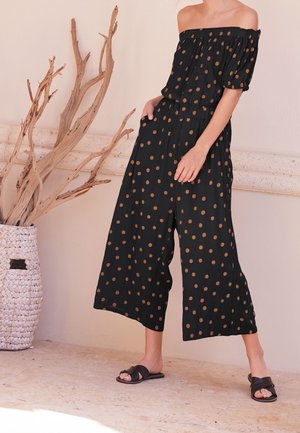 Sort off-shoulder jumpsuit med brune prikker, elastisk talje, løs pasform, cropped bukser, parret med sorte slide sandaler.