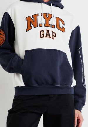Sudadera con capucha con sección superior blanca y sección inferior azul marino, mangas azules, texto naranja "N.Y.C GAP" en el pecho y un parche de baloncesto en el brazo izquierdo.