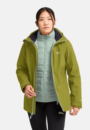 Woman wearing a light blue quilted jacket under an open olive green hooded raincoat, standing against a plain background.