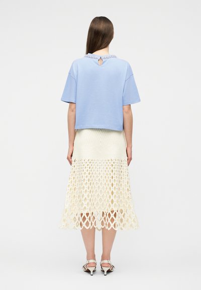 Γυναίκα με μακριά ίσια μαλλιά, φορώντας γαλάζιο κοντομάνικο τοπ και κρεμ μίντι φούστα με πλέξη κροσέ, μαζί με λευκές γόβες, με την πλάτη προς την κάμερα.