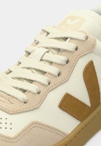 Κοντινό πλάνο κρεμ αθλητικού παπουτσιού με μπεζ σουέντ λεπτομέρειες, λευκά κορδόνια και καφέ λογότυπο "V" στο πλάι.