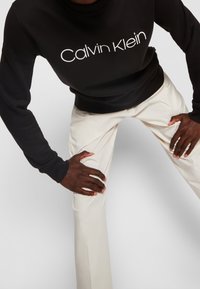 Zwarte katoen sweatshirt met het logo "Calvin Klein" in het wit, gecombineerd met crèmekleurige broek. Focus op handen die op de knieën rusten.