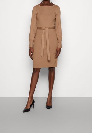 Kvinna i en beige långärmad klänning med bälte och svarta högklackade skor, stående mot en vit bakgrund.