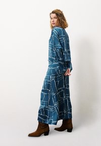 Lange, turquoise jurk van een zijdezacht materiaal, met een wit geometrisch patroon. Het ontwerp heeft lange mouwen en wordt gecombineerd met bruine enkellaarsjes.