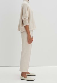 Suéter de punto beige con un cuello alto, corte relajado y hombros caídos, combinado con pantalones crema ajustados y zapatos de deslizar ligeros.