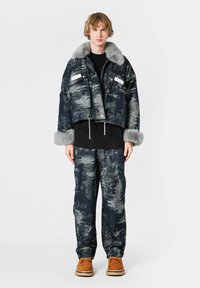 Chaqueta de denim cortada con cuello de piel sintética gris, dos bolsillos en el pecho, combinada con pantalones estampados en azul oscuro y gris, y calzado marrón.