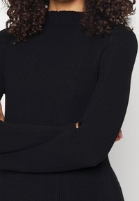 Maglione nero a coste con maniche lunghe e un alto collo scallop. Il tessuto presenta una superficie textured, evidenziando linee verticali.