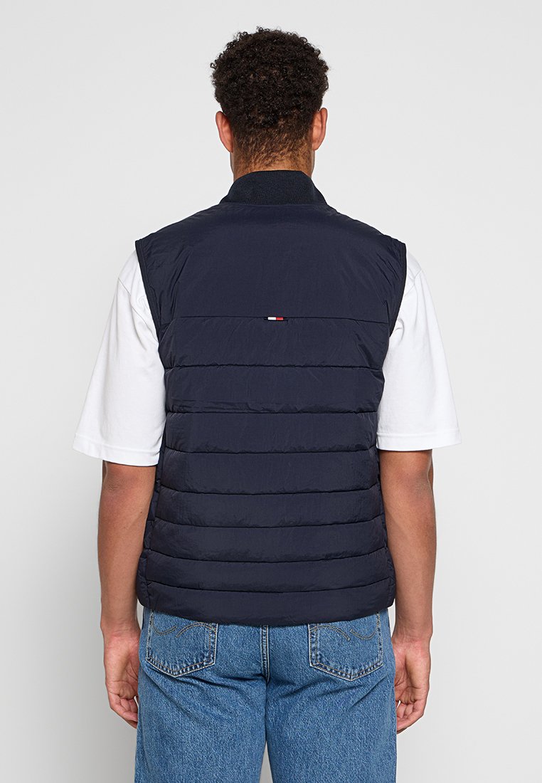 Chaleco tipo puffer azul marino con cuello alto, diseño acolchado horizontal en la parte delantera, detalle de logo en la parte trasera y textura de tela suave. Llevado sobre una camisa blanca.