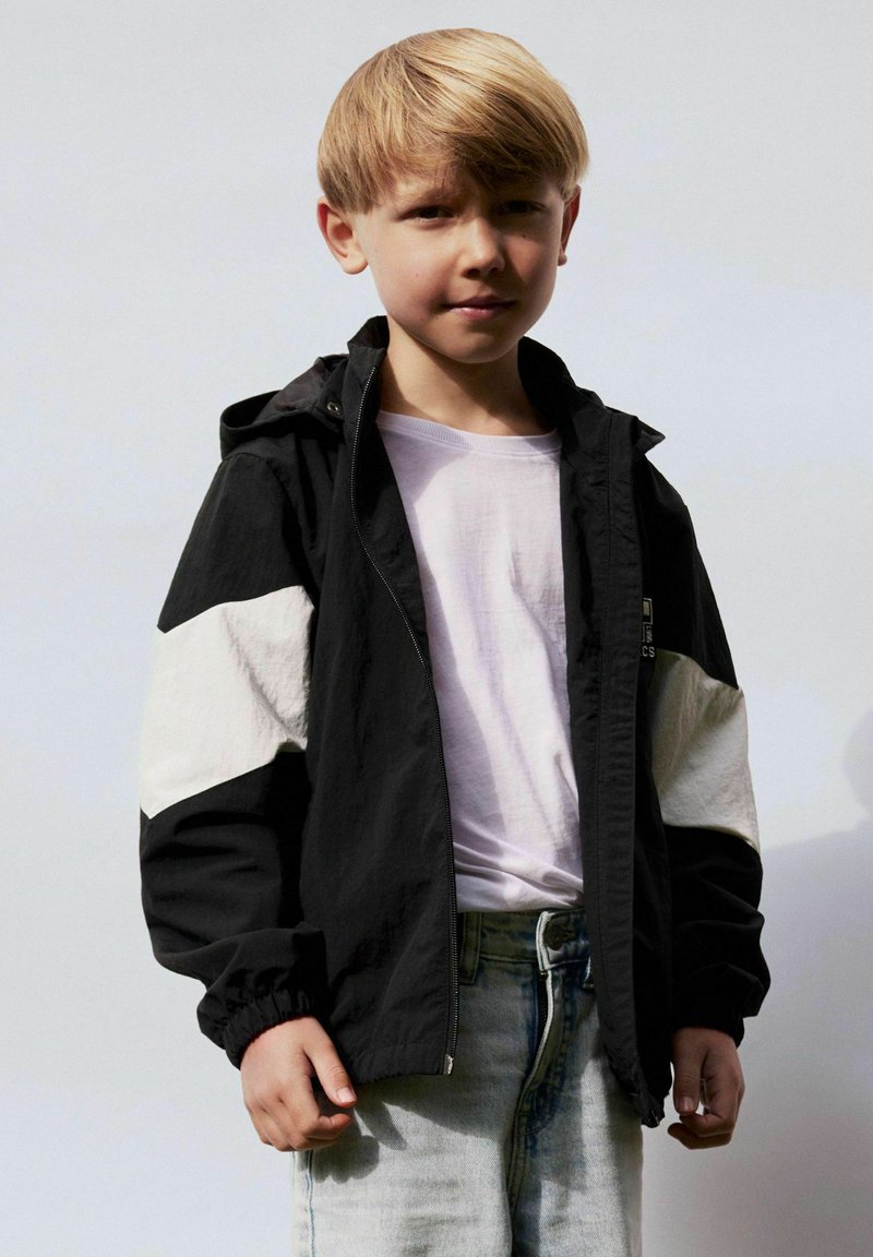 Ung dreng med blondt hår iført sort-hvid jakke over en hvid skjorte og lyseblå jeans, stående mod en ensfarvet lys baggrund.