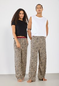 To modeller har på seg avslappede loungewear. Den ene har på seg en svart t-skjorte, mens den andre har på seg en hvit ermeløs topp. Begge har på seg bukser med leopardmønster og rød elastisk midje.