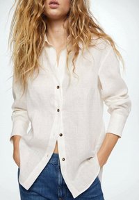 Kvinde med løst krøllet hår iført en hvid langærmet skjorte og blå jeans med hænderne i lommerne, stående foran en ensfarvet baggrund.