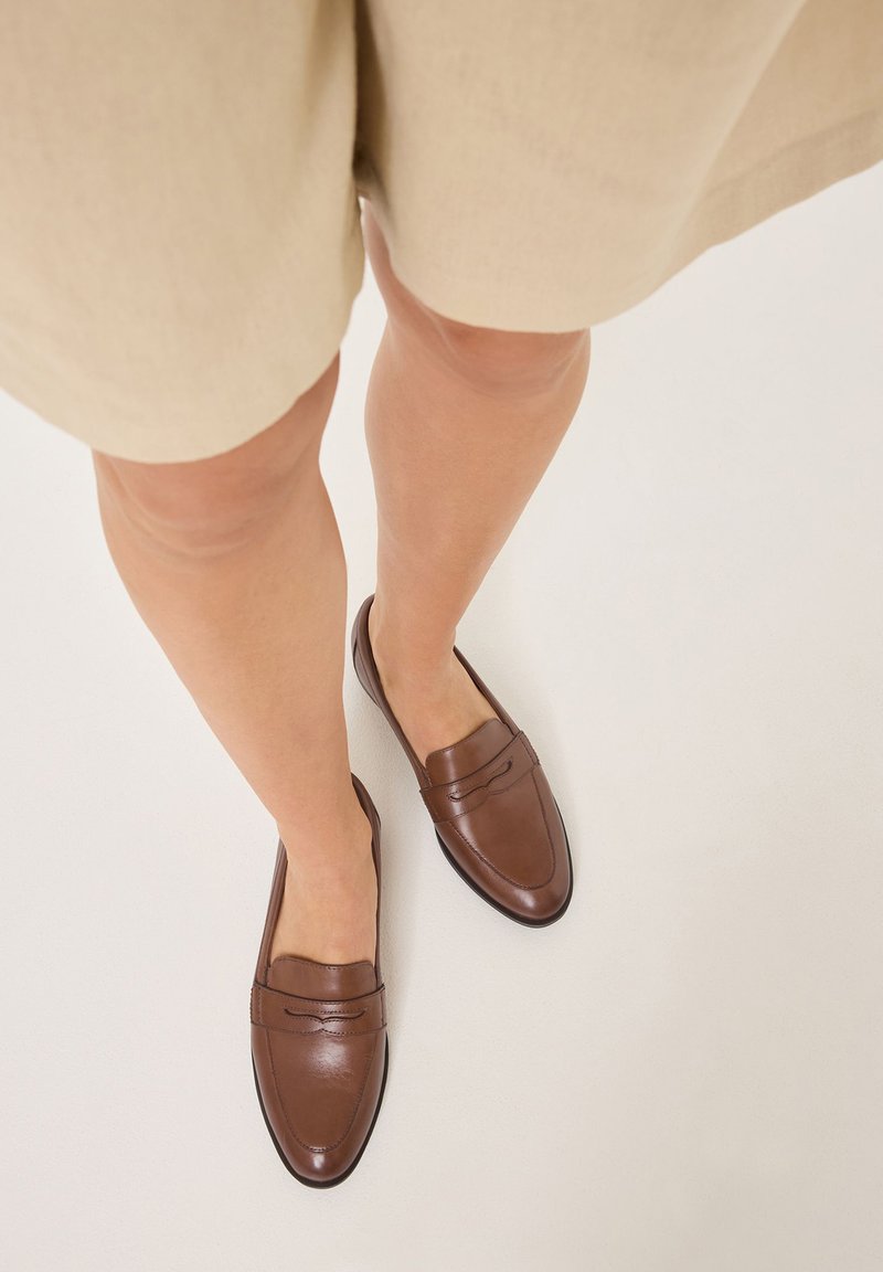 Braune Lederloafers mit glatter Textur, spitzem Zehenbereich und dekorativer Stickerei an der Vorderseite; kombiniert mit beigen Shorts.