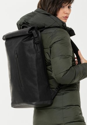 Person trägt eine olivgrüne Steppjacke und einen großen schwarzen Rolltop-Rucksack mit einer vorderen Reißverschlusstasche.