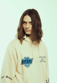 Sudadera beige con un gran gráfico azul que dice "Mud City REYES." Texto en las mangas: "Grime Infinito" y "Quema y Saqueo." Textura suave.