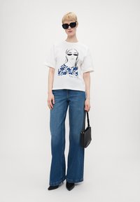 Hvit bomull t-skjorte med blå grafisk design. Vid ben jeans i blå. Svart håndveske med kort stropp. Svart katteøye solbriller.