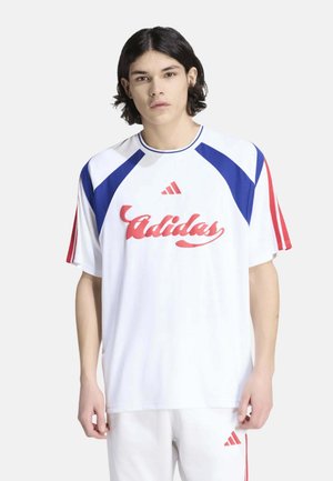 Joven con cabello hasta los hombros que lleva ropa deportiva blanca de Adidas con detalles en rojo y azul, de pie frente a un fondo liso.