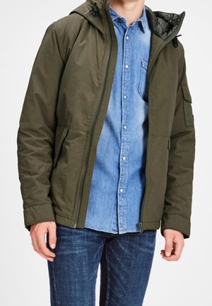 Mand iført olivengrøn hættetrøje over en lyseblå denimskjorte med knapper og mørkeblå jeans, stående med armene afslappede.