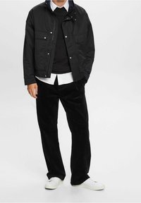 Mustat takki napin kiinnityksellä ja sivutaskuilla, kerrottu mustan villapuseron ja valkoisen paidan päällä. Mustat corduroy-housut ja valkoiset kengät täydentävät asukokonaisuuden.