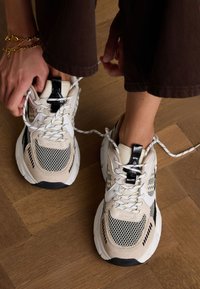 Beige und schwarze Netzsneaker mit weißen Schnürsenkeln, gepolsterter Zunge und strukturierten Akzenten. Dicke Sohle und gemusterte Details an den Seiten.