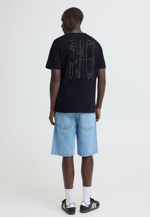 Homme portant un t-shirt noir avec des dessins techniques blancs dans le dos, un short en jean bleu clair, des chaussettes blanches et des baskets noires Adidas, vu de dos.