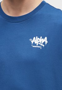 Μπλε μπλουζάκι με στρογγυλή λαιμόκοψη και λευκό λογότυπο «Alpha Industries» σε στυλ γκράφιτι τυπωμένο στο αριστερό μέρος του στήθους.