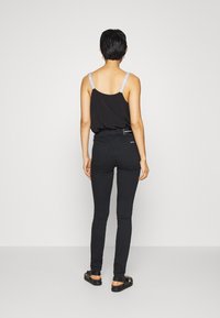 Top negro sin mangas con tirantes delgados y decorativos, combinado con jeans ajustados negros de cintura alta y sandalias planas negras. Fondo simple.