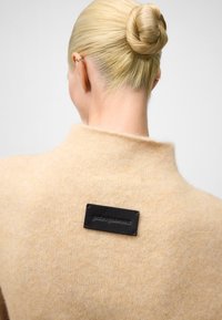 Pull beige en tissu doux avec un col haut, comportant une étiquette en cuir noir cousue à l'arrière. Texture lisse et design minimal.