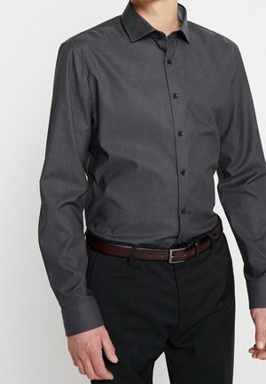 Homme portant une chemise cintrée gris foncé à boutons avec un motif subtil, un pantalon noir et une ceinture en cuir marron sur un fond uni.