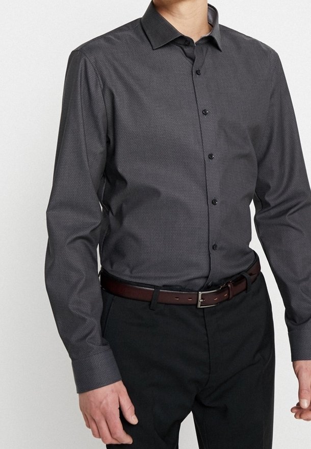 Homme portant une chemise cintrée gris foncé à boutons avec un motif subtil, un pantalon noir et une ceinture en cuir marron sur un fond uni.