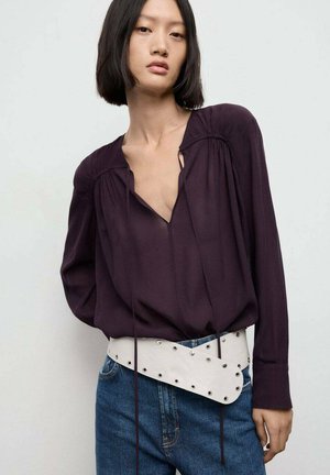 Mørk lilla langærmet bluse med dyb V-udskæring, præget af draperede detaljer. Sammen med et hvidt bælte, pyntet med sølvnitter, over et par blå jeans.