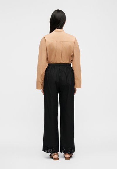 Γυναίκα με μακριά σκούρα μαλλιά, ντυμένη με μπεζ μπλούζα με μακριά μανίκια και μαύρο παντελόνι φαρδιά παντελόνα με δαντελένιο τελείωμα, στέκεται με την πλάτη προς τα πίσω σε λευκό φόντο.