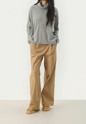 Femme portant un pull gris ample, un pantalon beige à jambes larges et des chaussures de style mocassin couleur tan, se tenant avec les mains dans les poches.