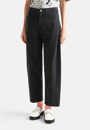 Person trägt schwarze High-Waist-Weitbein-Jeans, weiße Loafers, pinke Socken und ein graues Shirt mit Mickey-Mouse-Aufdruck, steht vor weißem Hintergrund.