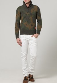 Strickpullover mit Tarnmuster in Grün und Braun, mit Kragen und Knopfdetails, kombiniert mit weißen Jeans und braunen Schnürschuhen.