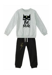 Grauer Hoodie mit schwarzem "BE BRAVE"-Text und fledermausähnlichem Design; kombiniert mit schwarzen Hosen, die ein gelbes Blitz-Patch haben.